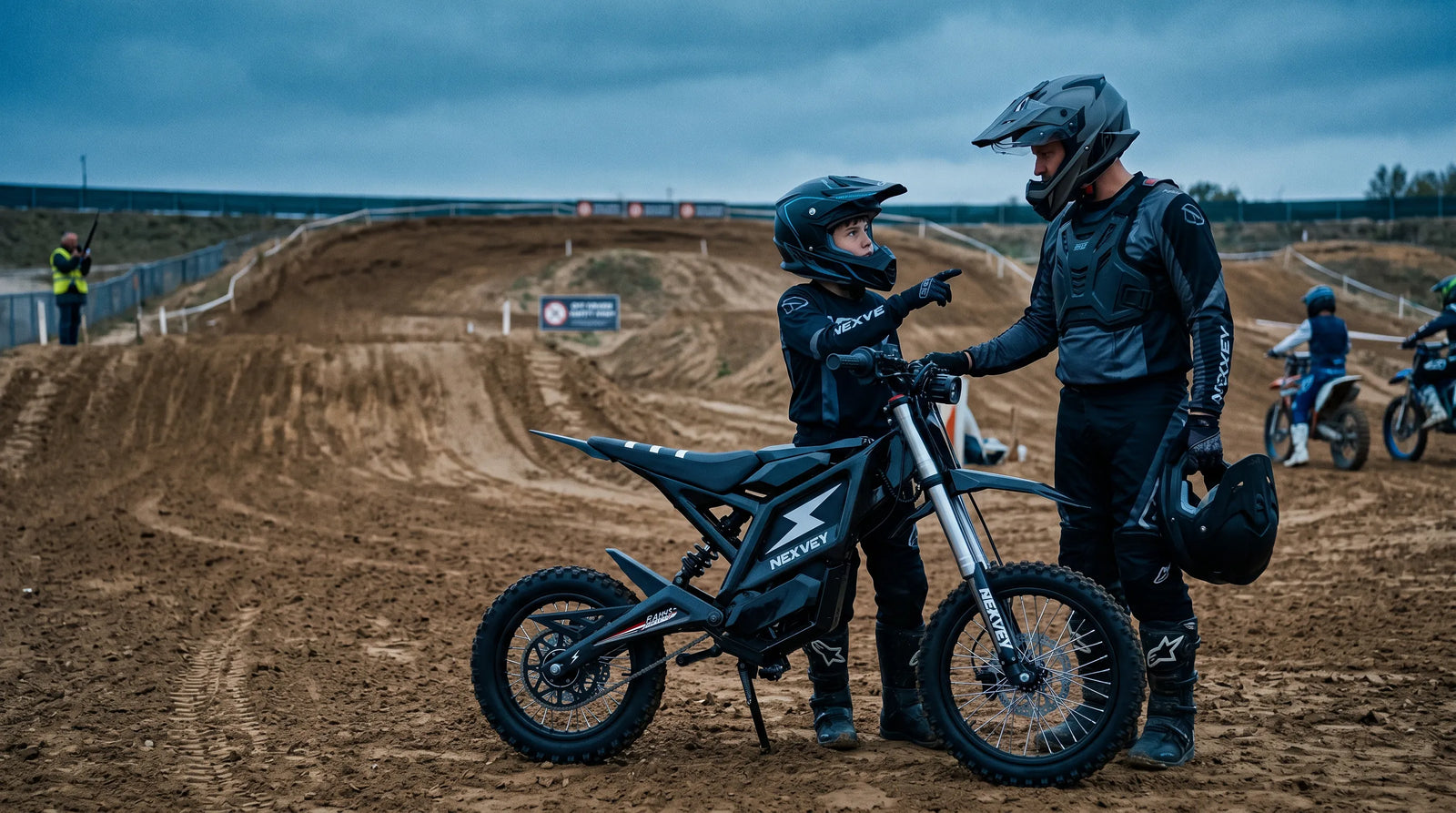   A teen wearing a full-face helmet stands beside an electric dirt bike with a parent. They are at a controlled off-road riding area; both look at the bike as if discussing safe riding.