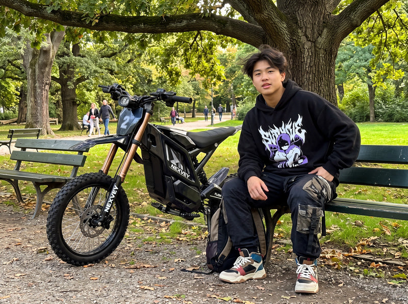 A rider resting on a park bench beside a black NEXVEY FLASH 2 electric dirt bike under a tree