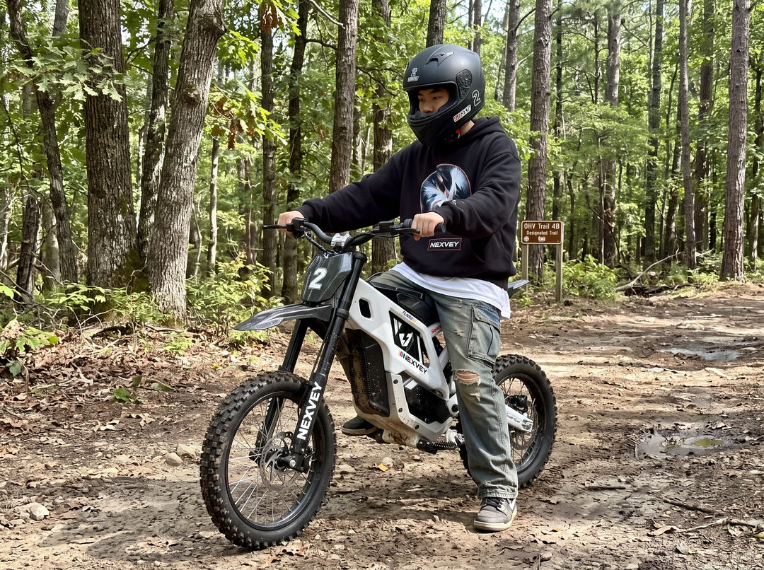 A helmeted rider in a black top on a white NEXVEY FLASH 2 electric dirt bike on a forest trail