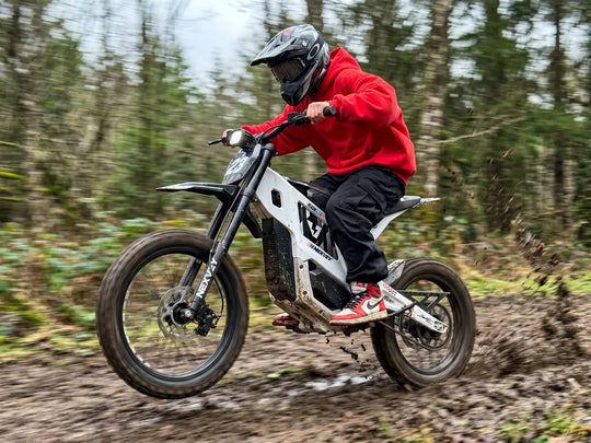 Rider in red hoodie riding the white Nexvey Flash Pro youth electric dirt bike through a muddy off-road forest trail