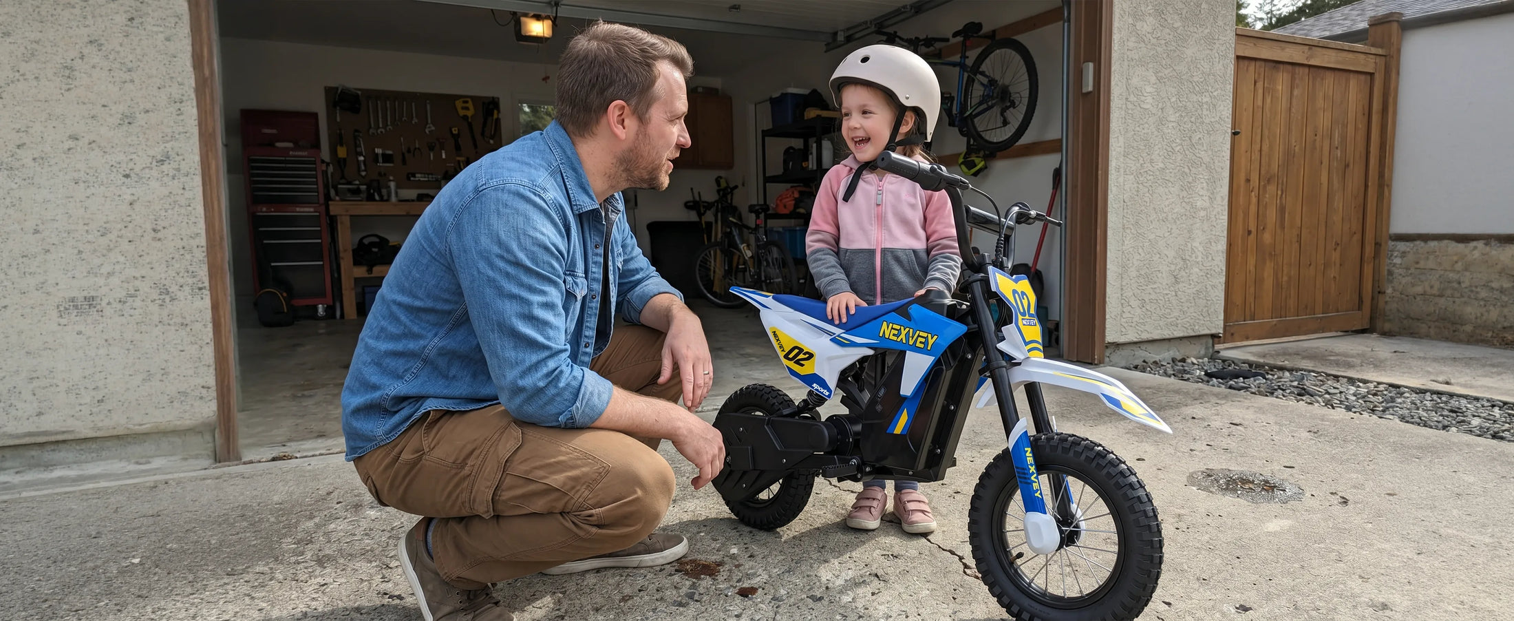 Child wearing a helmet sitting on a blue NEXVEY Spark 02 electric dirt bike with a parent in front of a garage