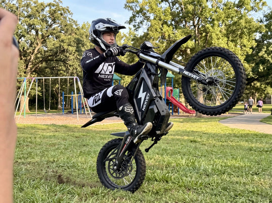 A young rider in professional racing gear performing a high-angle wheelie on a black NEXVEY YOUTH electric dirt bike on the grass at a local park playground