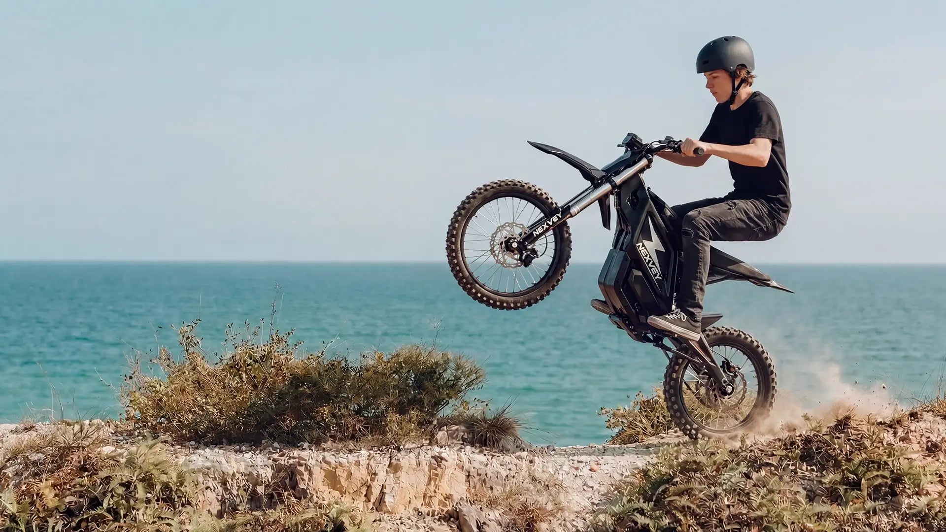 A rider performing a wheelie on a NEXVEY electric dirt bike on a coastal cliff with the ocean in the background